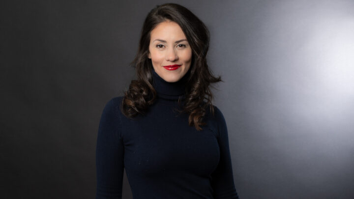 A global compliance manager with long dark hair and red lipstick is wearing a black turtleneck sweater, standing against a dark gray background, ready to address the team at Sermo.