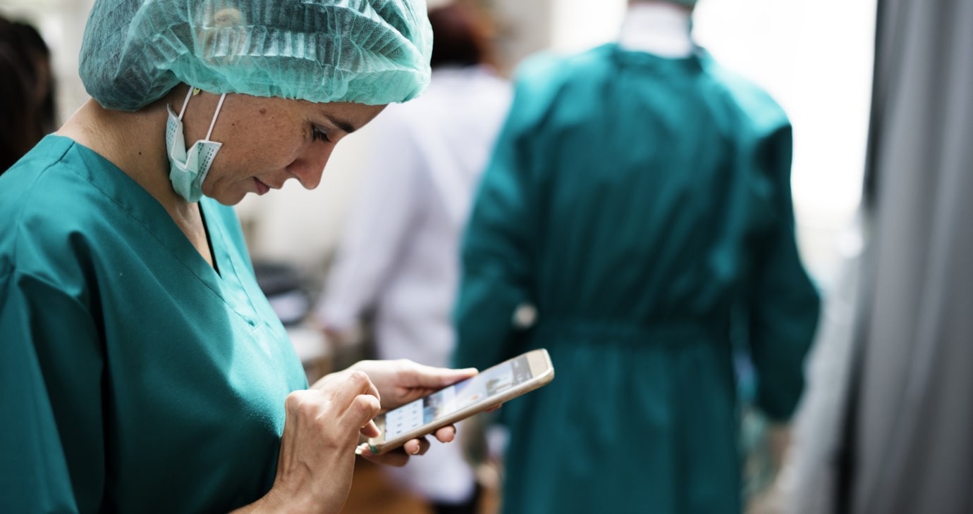 A medical professional in scrubs and a hair cover uses a smartphone, possibly utilising AI for medical diagnosis. Two more individuals in medical attire stand in the background, contributing to this innovative approach in predicting patient outcomes.