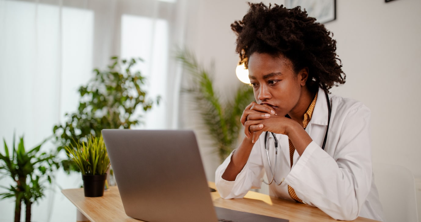 A focused individual in a white lab coat sits at a desk with a laptop, hands clasped near chin, surrounded by plants in a brightly lit room. The setting pulses with the promise of medical AI, predicting patient outcomes to revolutionise healthcare.