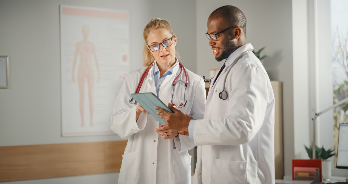 Two doctors in white coats discuss information on a tablet utilising medical AI in their office. A poster of the human anatomy is visible in the background, underscoring how AI for medical diagnosis is revolutionising patient care.