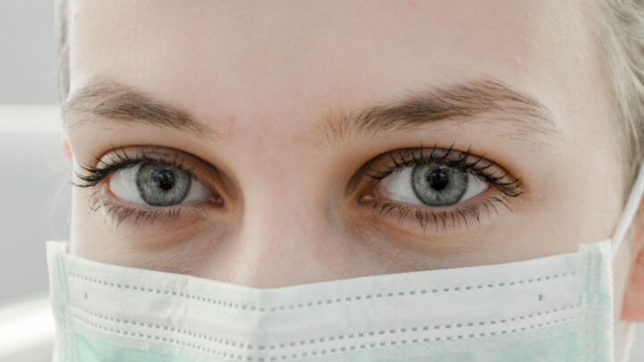A close-up captures a person wearing a light green surgical mask, reflecting the ongoing medical shortages during COVID-19. Their blue eyes peek over the mask with a neutral expression, revealing resilience shared by physicians worldwide.