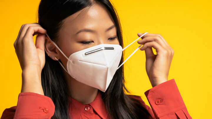 A person in a red shirt carefully lifts their white face mask against a vibrant yellow background, resembling the attentive gesture of doctors.
