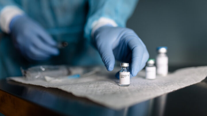 A medical professional in blue gloves carefully reaches for a Johnson & Johnson COVID-19 vaccine vial on a table with more vials and a syringe.