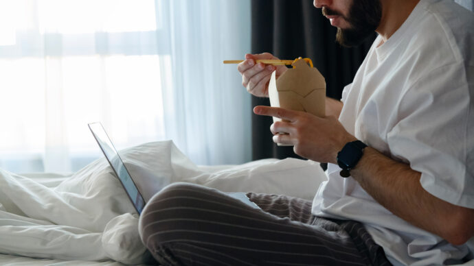 A person in pajamas is sitting on a bed, using chopsticks to eat from a takeout container while looking at a laptop, reflecting on pandemic weight amidst the comfort of cozy routines.