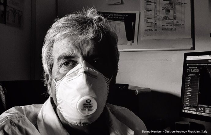 A grayscale image captures a person wearing a white face mask, likely due to the coronavirus, seated in front of a computer screen displaying data, while a bulletin board looms in the background.