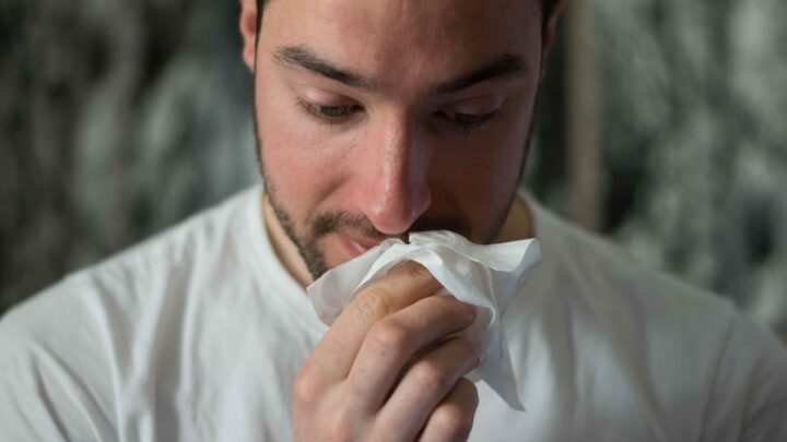 A man in a white shirt holds a tissue to his nose, appearing to sneeze or wipe his nose, perhaps due to the pesky influenza virus that often strikes during winter.