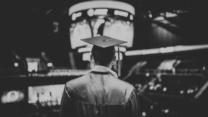 A med student in a cap and gown stands facing a large screen in a dimly lit arena, reflecting on graduating early during the COVID-19 pandemic.