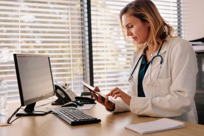 A physician in a white coat and stethoscope sits at a desk, using a tablet. There is a computer, phone, and notepad on the desk—essential tools for managing various content types. Blinds cover the window in the background, adding to the focused atmosphere.