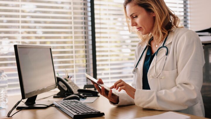 A physician in a white coat and stethoscope sits at a desk, using a tablet. There is a computer, phone, and notepad on the desk—essential tools for managing various content types. Blinds cover the window in the background, adding to the focused atmosphere.
