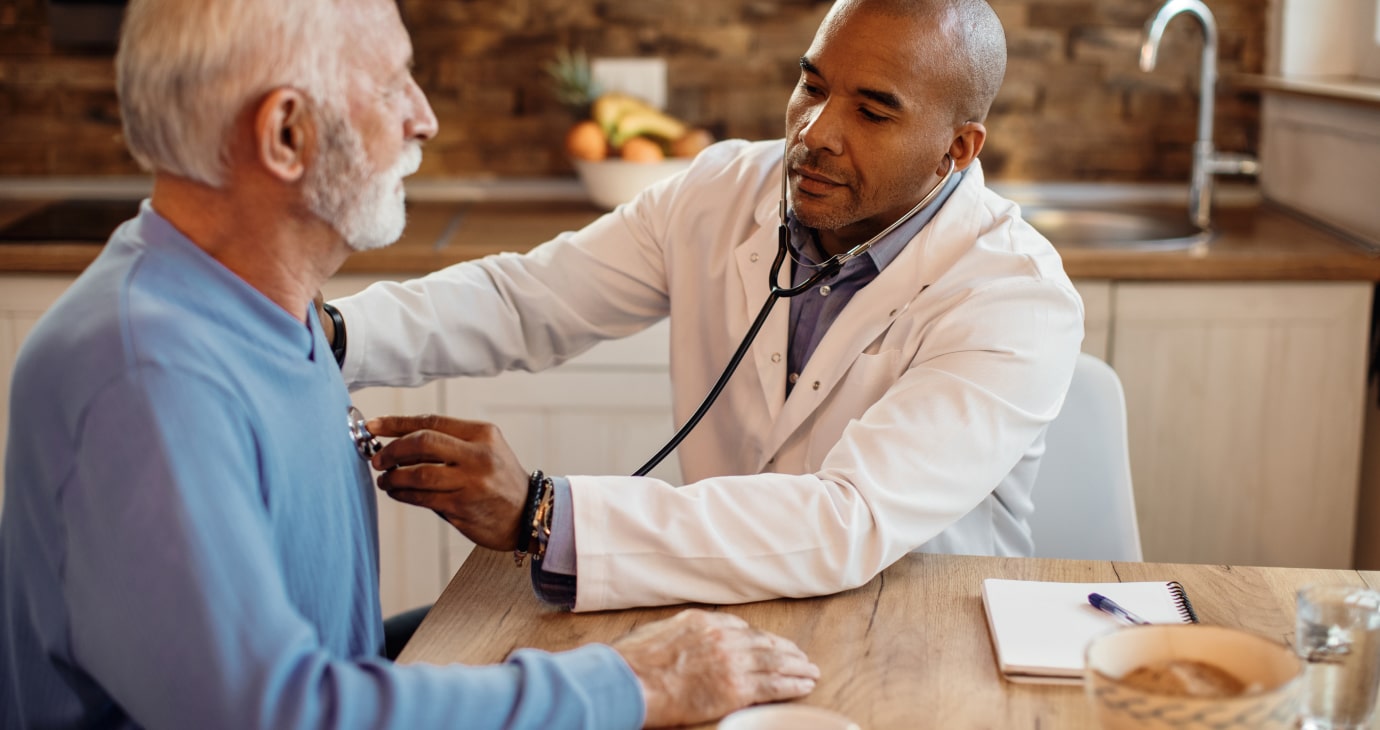 A physician uses a stethoscope to examine an elderly man in a casual setting, with a notebook and a plant in the background, ensuring patient satisfaction.