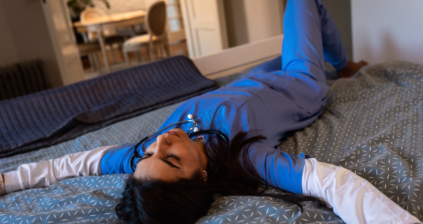 Una doctora en pijama sanitario azul se relaja sobre una cama, con los brazos extendidos y el fonendo colgado al cuello, en un momento de paz en medio de una crisis de salud mental.