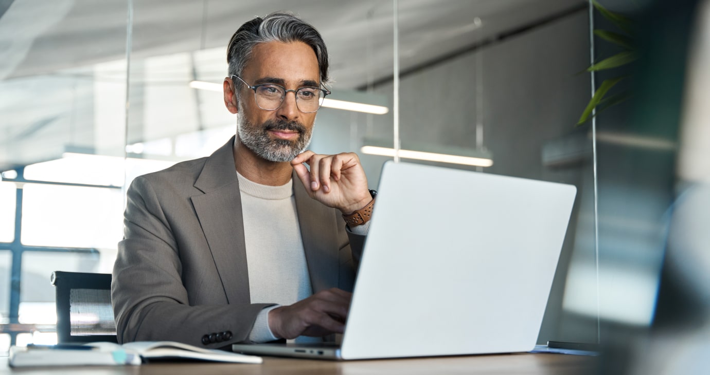 Un hombre con gafas y barba está sentado en un escritorio, usando un ordenador portátil. Lleva una chaqueta beis y mira la pantalla con atención, tal vez estudiando las últimas reflexiones sobre la salud mental.