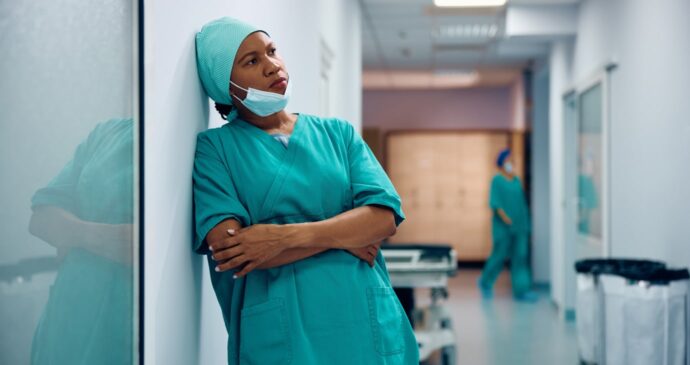 A healthcare worker in green scrubs and cap leans against a wall in a hospital corridor, contemplating the ethical perspectives of assisted dying. Another person is visible in the background.