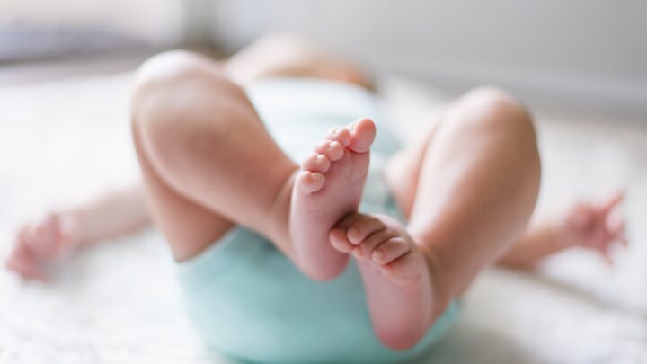 A baby, nestled on their back in a light blue outfit with tiny feet playfully in the air on a soft, white surface, embodies the joy of early days while underscoring physicians' concern amid the decline in childhood vaccinations.