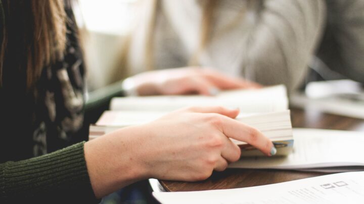 Two people sit at a table with open books, immersed in the world of higher education. One person's hand is focused on turning a page, while papers are scattered across the table, reminiscent of a time when pandemic-driven challenges shaped their academic journey.