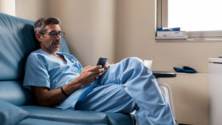 A credible healthcare practitioner in blue scrubs and glasses sits on a couch, using their smartphone near a window, possibly engaging with social channels.