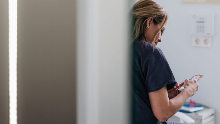 A woman in a navy blue shirt is sitting and looking at her phone with her head slightly turned, optimizing her social media strategy. The background features a white wall and some office equipment, creating the perfect setting for focused planning.