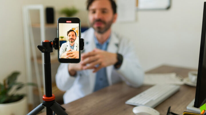 A doctor in a white coat is talking on a video call, like an influencer, using a smartphone mounted on a tripod.
