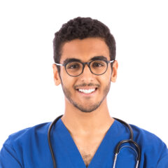 Una persona con cabello corto y rizado y gafas, que viste un uniforme médico azul y un estetoscopio, sonríe a la cámara, reflexionando sobre las diversas ocupaciones secundarias para médicos.