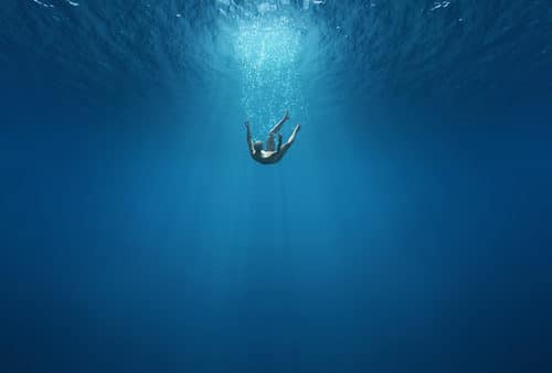 A person floats serenely underwater, surrounded by the deep blue ocean with sunlight filtering through, creating an atmosphere so tranquil that it feels as if they are momentarily drowning in peace.