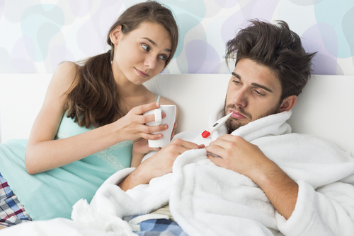 A woman compassionately offers a mug to a man suffering from the dreaded man flu, sitting on the bed in his bathrobe, clutching a thermometer.
