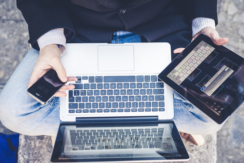 A person sits cross-legged, seamlessly multitasking with a smartphone and tablet in hand while efficiently using a laptop on their lap.