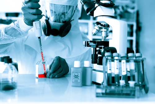 In protective gear, a scientist carefully uses a pipette in a lab filled with microscopes and test tubes, aware of the potential risk involved in synthesizing viruses.