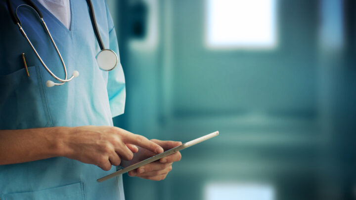 A healthcare professional in scrubs uses a tablet with a stethoscope around their neck in a clinical setting, tapping into the power of medical crowdsourcing. Through platforms like Sermo, solutions are solved collaboratively by connecting with experts worldwide.