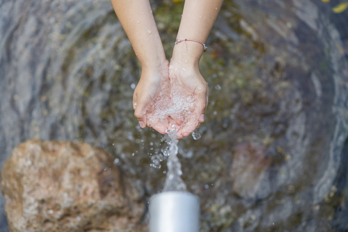 Hands cupping raw water flowing steadily from a pipe above a rugged, rocky surface.