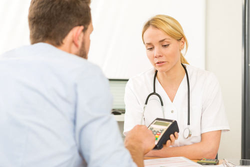 A healthcare professional with a stethoscope offers software advice while discussing a handheld device with a man in a medical setting.