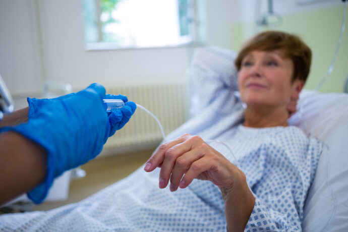 A patient in a hospital bed receiving IV treatment, with someone in blue gloves adjusting the drip as they monitor symptoms.