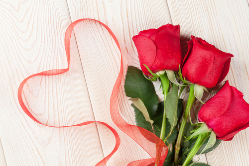 Three red roses with green leaves lie on a wooden surface, accompanied by a red ribbon shaped like a heart—a perfect valentine's arrangement.