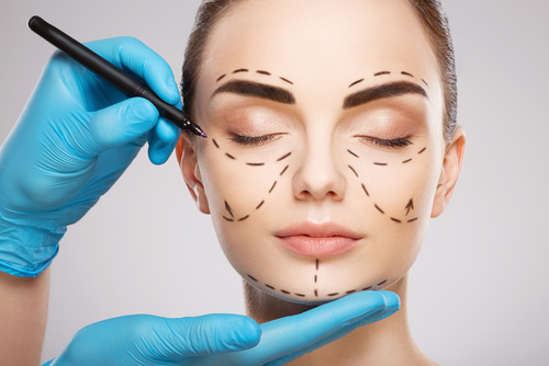 A person with closed eyes has facial markings drawn with dashed lines, hinting at a plastic surgery procedure. A gloved hand supports their chin while another holds a marker near their forehead.