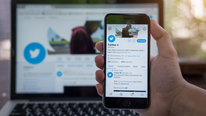 Close-up of a hand holding a smartphone displaying a Twitter profile, highlighting the triumphs of independent practice. In the background, the same profile is open on a laptop screen.