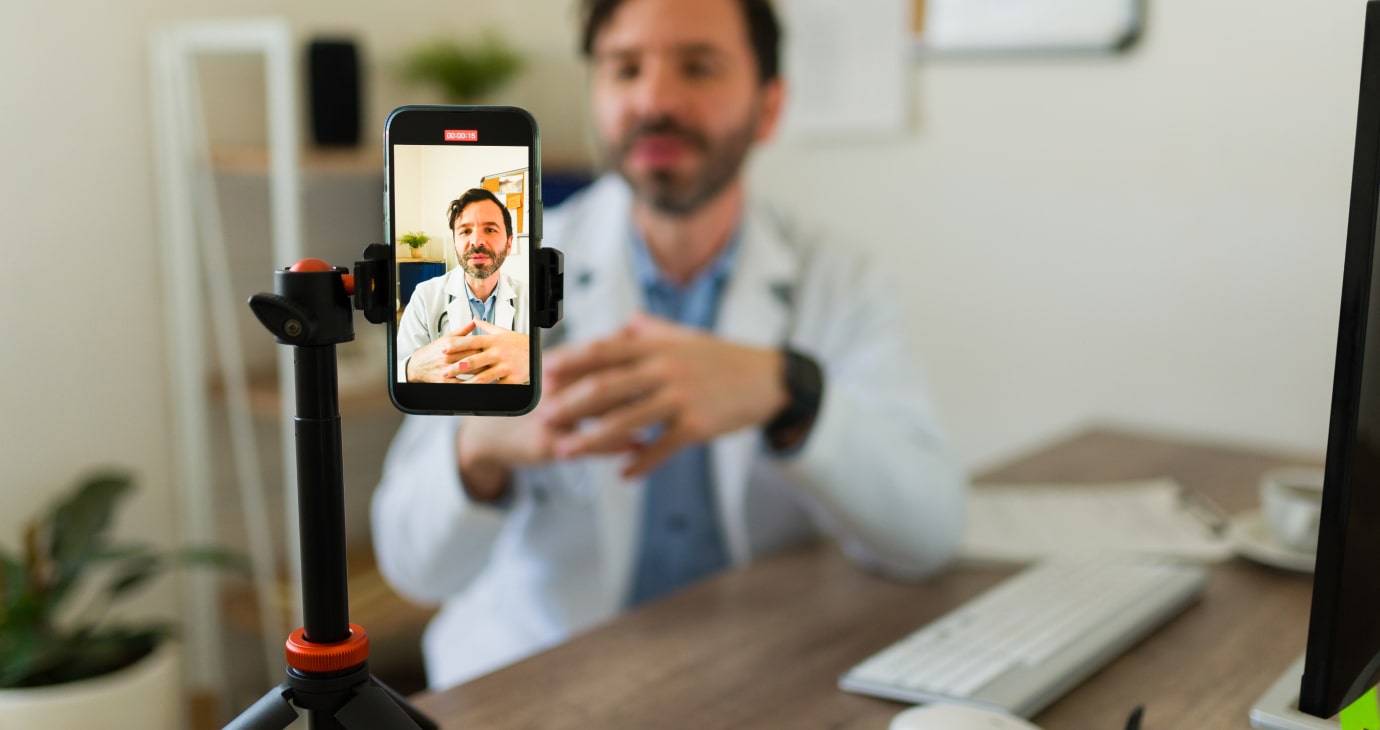 Un médico con bata blanca graba un vídeo para TikTok en un teléfono móvil montado en un trípode, exhibiendo experiencia sanitaria en un entorno de consulta.