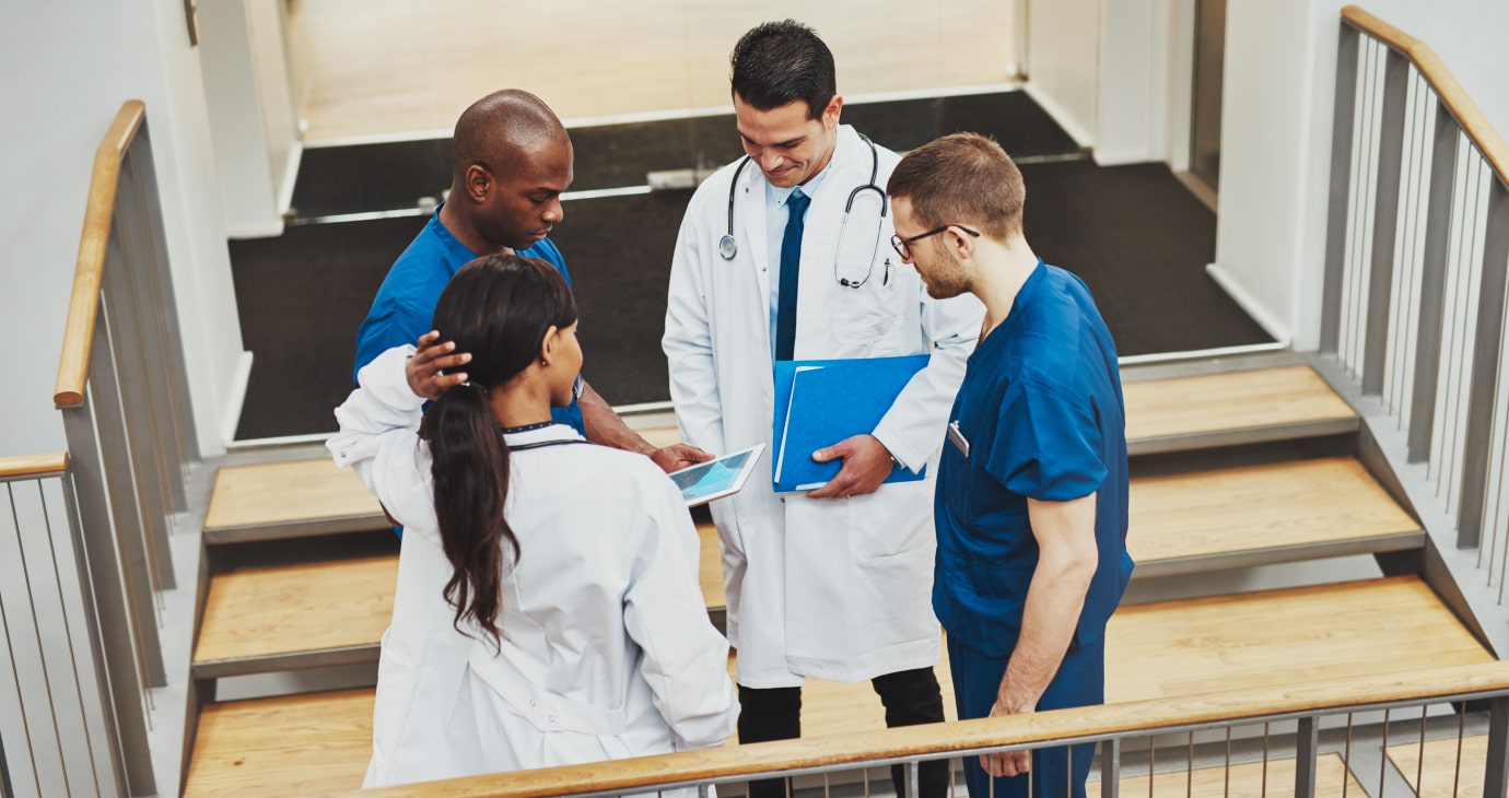 Cuatro profesionales de la salud, probablemente médicos destacados en TikTok, conversan en una escalera. Dos en bata blanca y los otros dos en uniforme quirúrgico azul, uno tiene una tablet mientras que otro tiene una carpeta azul en la mano, posiblemente sopesando su próxima publicación viral en las redes sociales.