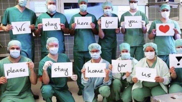 A group of medical staff in scrubs and masks, representing the physician community, hold signs saying "We stay here for you. Please stay home for us" with a heart symbol, highlighting their dedication during COVID-19.