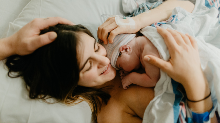 Mother, once pregnant, now lies in bed holding her newborn baby on her chest, with a hand gently touching her head.