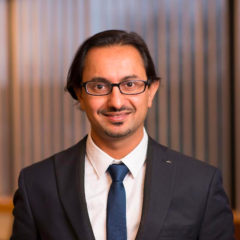 El Dr. Shivang Joshi, un estimado MPH y MD, se mantiene firme con traje y corbata, mientras sus anteojos reflejan la luz de las persianas verticales detrás de él.