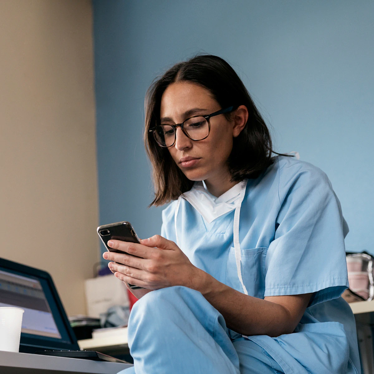 Una persona con uniforme y gafas está sentada, mirando un teléfono, con una computadora portátil cerca, contra una pared azul.